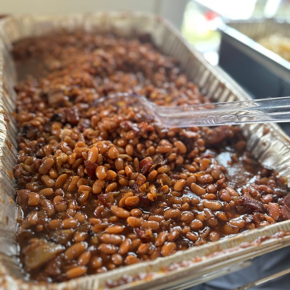 A large foil pan filled with baked beans, glistening with sauce, and a serving spoon.