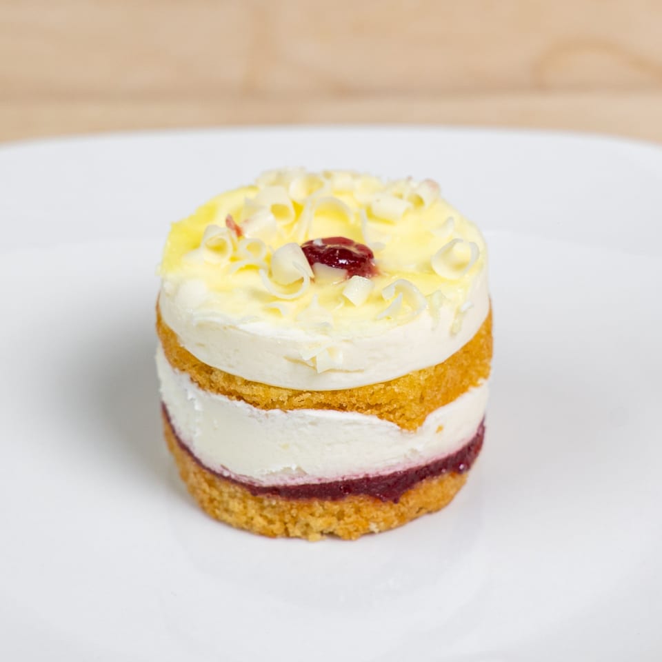 A layered dessert with yellow cake, white cream, and red jam, topped with white chocolate curls.
