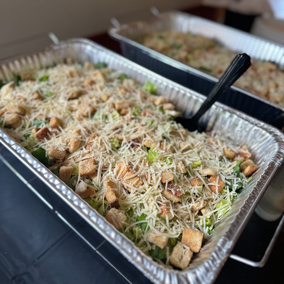 Two large catering pans of Caesar salad with croutons and shredded cheese, ready to serve.